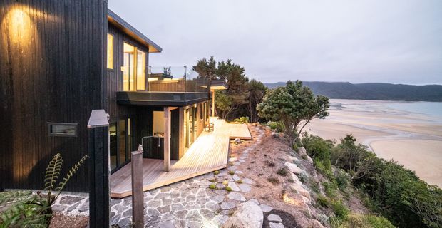 A contemporary multi-level coastal house set on a steep, elevated site above a broad sandy beach and tidal estuary. The dark timber-clad facade, extensive glazing, cantilevered upper level and warm timber deck are visible, with stone paving and native coastal vegetation surrounding the building.