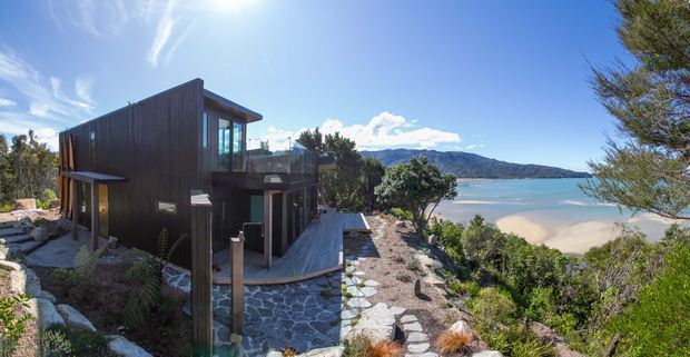 Contemporary multi-level dark-clad coastal residence set on a rocky, landscaped hillside overlooking a sandy beach, calm blue water and mountainous shoreline. The home features expansive glazing, projecting roof forms, timber decking, stone retaining walls and a stepping-stone path.