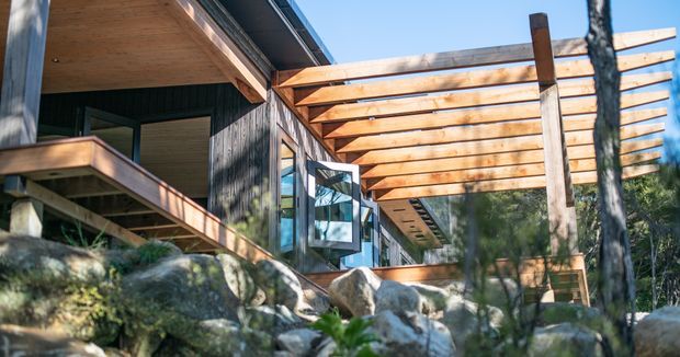 Exterior view of a contemporary timber-clad house set among rocky natural terrain and trees. The image highlights a broad timber pergola and covered deck with exposed beams, timber soffits, large glazed openings, and a projecting upper roofline.