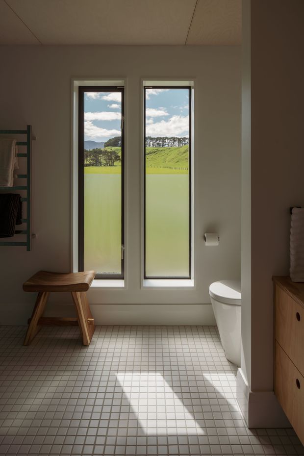 A bright, minimalist bathroom with two tall narrow windows overlooking a green rural landscape. The room features small square light floor tiles with dark grout, pale walls and ceiling, a white toilet, timber vanity cabinetry, and a simple timber stool beside a dark towel rail. Sunlight creates strong geometric shadows across the floor.