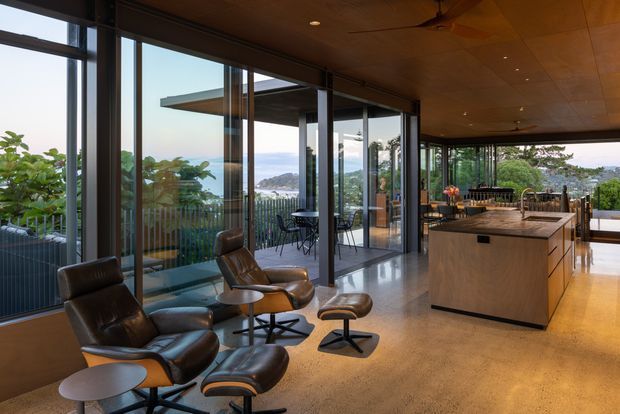 A spacious, refined open-plan living and kitchen interior with floor-to-ceiling glazing overlooking a leafy, hilly landscape and water. The room features a warm timber ceiling, polished concrete flooring, a large timber kitchen island, dark-framed sliding glass doors, and a pair of leather lounge chairs with ottomans. A dining area and outdoor terrace are visible beyond the kitchen.