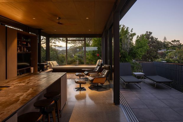 Warm, mid-century-inspired open-plan living and entertaining space with a stone-topped kitchen island, timber built-in shelving, leather lounge chairs, polished concrete flooring, and a timber-lined ceiling. Full-height glazed sliding doors open directly to a tiled outdoor terrace with dark balustrading and outdoor lounge furniture, overlooking mature vegetation.