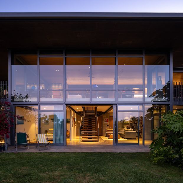 Twilight exterior view of a contemporary multi-level home with a broad glazed facade, dark horizontal roof overhang, and full-height aluminium-framed windows and doors. The transparent facade reveals a warmly lit interior with a central open-riser staircase, living area, and upper-level spaces. A lawn and planted garden border the building.