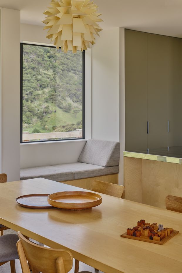 A warm, refined dining area with a large light-wood dining table and sculptural wooden chairs. A built-in upholstered window seat overlooks a green hillside, while tall dark olive cabinetry and a timber-fronted counter occupy the right side. A striking layered pendant light hangs above the table, with simple wooden trays and a board game as tabletop accents.