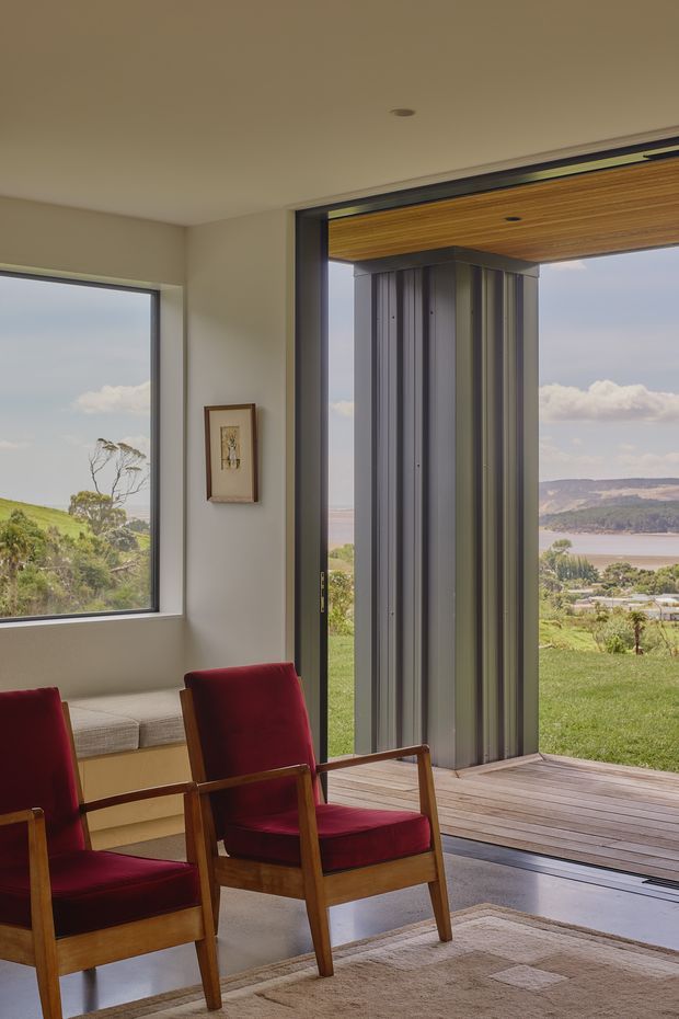 A calm, light-filled living room opening onto a timber deck, with large windows and a wide glazed doorway framing a grassy landscape, water, and hills beyond. The room contains two mid-century-style wooden armchairs upholstered in deep red, a built-in window seat, framed artwork, a neutral rug, and a timber-lined exterior soffit. A vertically ribbed dark metal-clad structural column is visible at the opening.