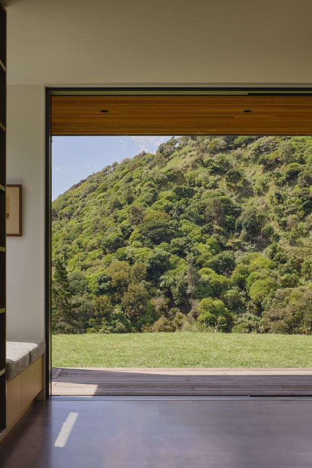 A minimalist interior looking through a large open or fully glazed opening toward a lush, steeply wooded hillside. The room has smooth light walls, a polished grey concrete floor, a dark-framed opening, a timber-lined overhead panel, and a timber deck outside. A partial built-in bed or bench is visible at the left edge.
