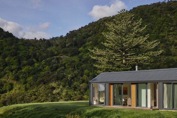 A low, modern single-storey house with extensive floor-to-ceiling glazing and a dark metal pitched roof, set in a grassy clearing against a densely forested hillside. Warm timber-framed glazed doors and neutral exterior panels create a simple connection to the surrounding landscape.