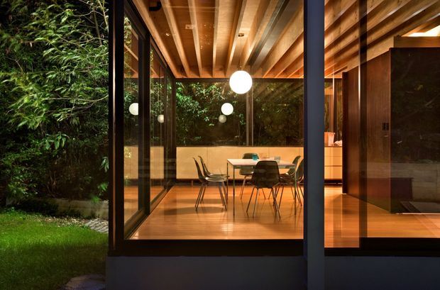 A warmly lit contemporary dining area viewed through expansive floor-to-ceiling glazing, opening onto a lush garden. The room features a timber-lined ceiling with exposed beams, timber flooring, a dark-framed glazed facade, a dining table with modern chairs, spherical pendant lights, and built-in timber cabinetry. A dark corridor or adjoining interior space is visible to the right.