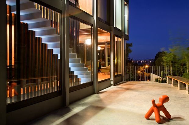 Night-time view of a contemporary multi-level residence opening onto a broad concrete terrace. Floor-to-ceiling glazing reveals a sculptural floating staircase, warm timber-lined interior surfaces, and modern furnishings. The terrace has a simple metal balustrade and bench seating, with a brightly coloured sculptural outdoor object in the foreground. The composition highlights the glazed facade, indoor-outdoor connection, and dramatic evening lighting.