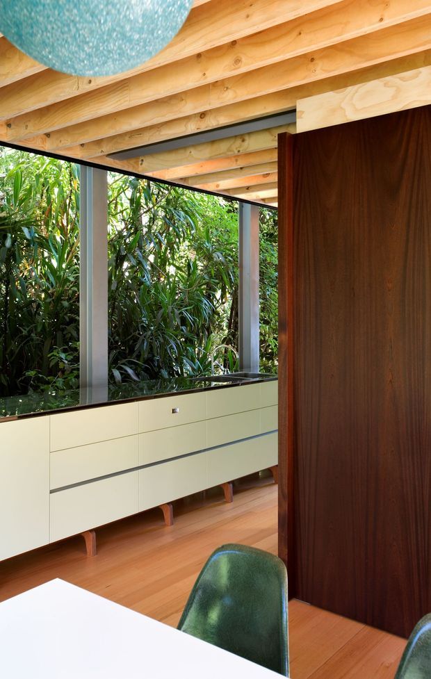 A warm, timber-lined open-plan interior with exposed wood ceiling beams, a long pale built-in sideboard, and a dark timber partition or cabinet. Full-height glazing opens visually onto dense greenery, while green upholstered dining chairs and a white tabletop appear in the foreground. The space has a strong connection to nature and abundant natural light.