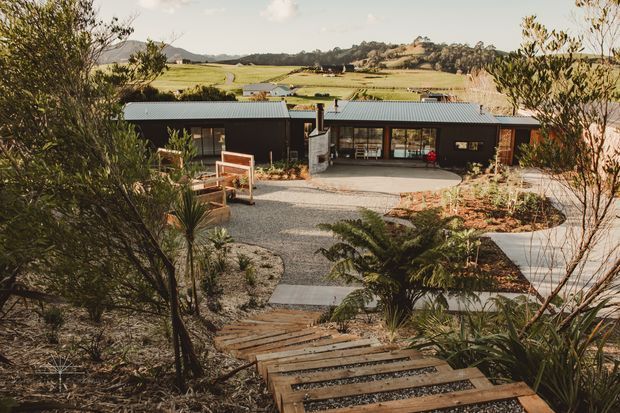 A low, contemporary rural home viewed from a landscaped hillside garden. The dark-clad single-storey building has long grey metal pitched roof planes, extensive glazing, and a covered central entry or carport-like recess. A gravel forecourt, timber stepping walkway, native-style planting, and established fern-like vegetation lead toward the house, with open pastoral farmland and rolling hills beyond.