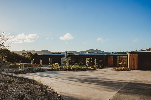 Wide exterior view of a low-slung contemporary single-storey residence with dark cladding, timber-toned garage or entry doors, extensive concrete driveway and landscaped planting. The home has a largely flat roofline and broad glazed openings, with rolling hills visible in the background under a clear blue sky.