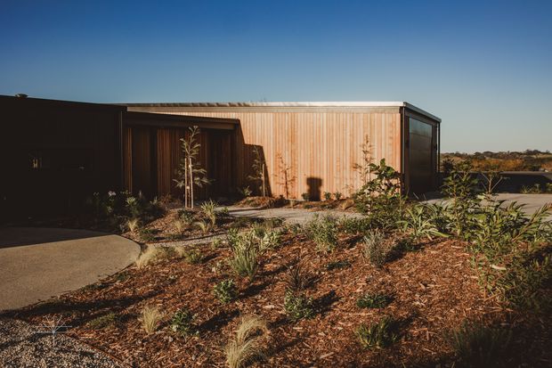 Single-storey contemporary flat-roof house set within a dry, landscaped site. The exterior combines dark cladding at the entry with warm vertical timber cladding on the main rectangular volume, framed by dark glazing and a slim light metal roof edge. Native-style low planting and mulched garden beds surround a curved concrete path, with water or open landscape visible beyond the building.