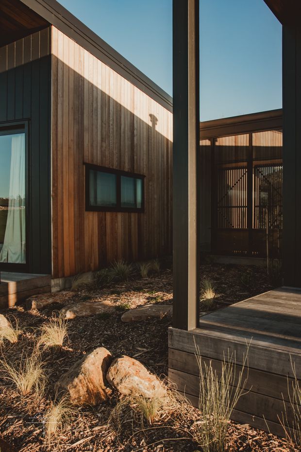 Exterior architectural view of a compact timber-clad building arranged around a landscaped courtyard. Vertical natural timber boards, dark structural posts and trims, large glazed openings, and a covered timber deck frame a sheltered entry or outdoor living area. Dry grasses, rocks, mulch, and dappled low-angle sunlight create a warm, restrained natural setting.