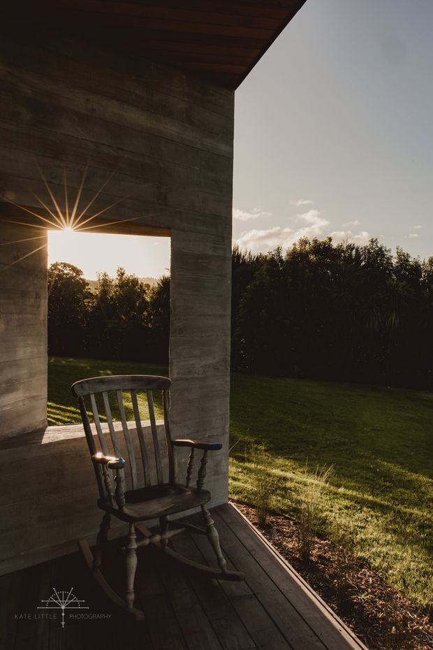 A moody architectural photograph of a covered outdoor porch or terrace framed by thick dark concrete walls and a timber-lined soffit. A traditional wooden rocking chair sits on dark timber decking beside a large rectangular opening, looking out toward a sunlit lawn and dense garden planting. Low golden sunlight creates a strong starburst through the opening, contrasting with the shaded structure.