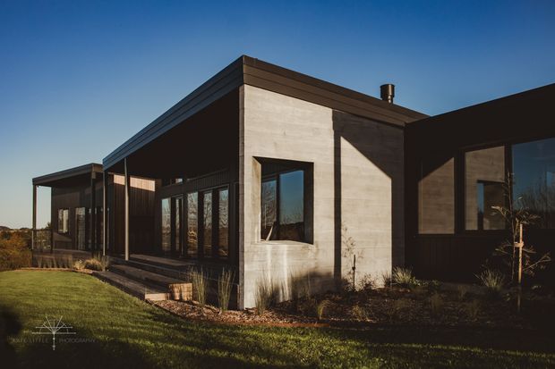 Single-storey modern residence with a low, angular flat-roofed form, dark cladding, extensive floor-to-ceiling glazing, and a prominent raw concrete wall volume. The house sits in a landscaped suburban or rural setting under clear evening light, with strong geometric shadows and restrained detailing.