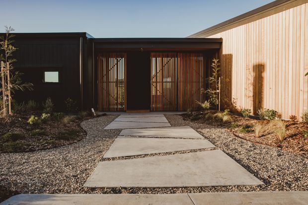 A single-storey modern building entrance viewed from the front, with dark charcoal cladding, a warm vertical timber-clad wall, and a recessed entry framed by vertical timber battens. Large rectangular concrete stepping slabs form a path through a gravel and drought-tolerant landscaped forecourt. The composition is minimalist and strongly geometric, with warm low-angle sunlight creating long shadows.