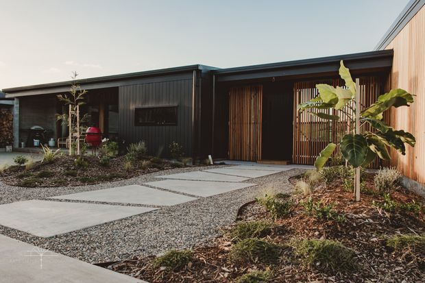 Single-storey contemporary house exterior with a low-pitched roof, dark vertical cladding, warm timber screening and timber wall cladding. A gravelled front landscape includes large concrete stepping slabs, native-style planting, and a recessed entry behind vertical timber battens.
