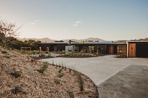 Wide exterior view of a low, single-storey contemporary home set within a landscaped, drought-tolerant garden. The dark horizontal facade has extensive glazing, timber-clad doors and screens, and a broad concrete driveway or forecourt. Rolling hills and a softly lit sky form the distant background.