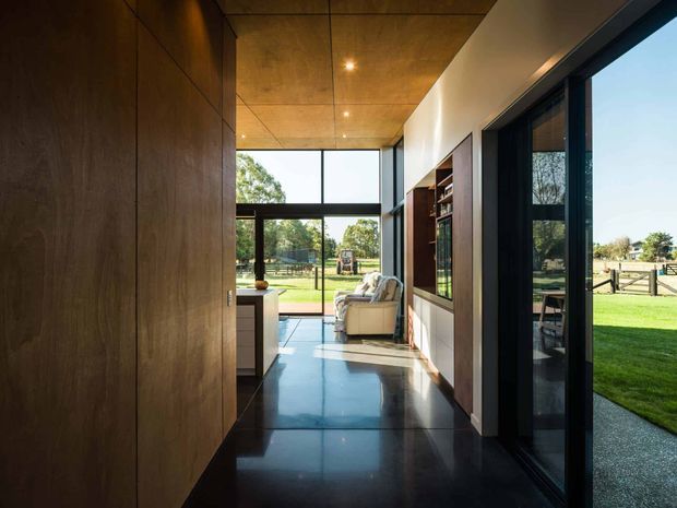 A refined contemporary residential interior hallway or circulation space with dark timber wall panelling and cabinetry, polished dark flooring, and a warm timber-lined ceiling with recessed spotlights. Floor-to-ceiling black-framed glazing opens the corridor visually to a green rural landscape, while built-in shelving and a seating area are visible further along. The space has strong indoor-outdoor connection, clean linear detailing, and abundant natural light.