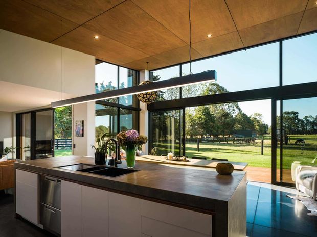 A contemporary open-plan kitchen centred on a large island with a dark stone benchtop, integrated sink and stainless-steel appliances. Full-height black-framed glazing opens the room to a green rural landscape and a blue-toned outdoor area. The space features a warm timber-panelled ceiling, white built-in cabinetry, a black linear pendant light and polished dark flooring.
