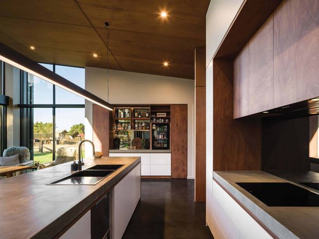 Contemporary kitchen with a large island, integrated sink and tap, dark stone-look benchtops, white lower cabinetry, timber veneer cabinetry and a built-in drinks display at the rear. The room features expansive black-framed glazing, a warm timber ceiling, dark flooring, recessed lighting and a linear pendant above the island.