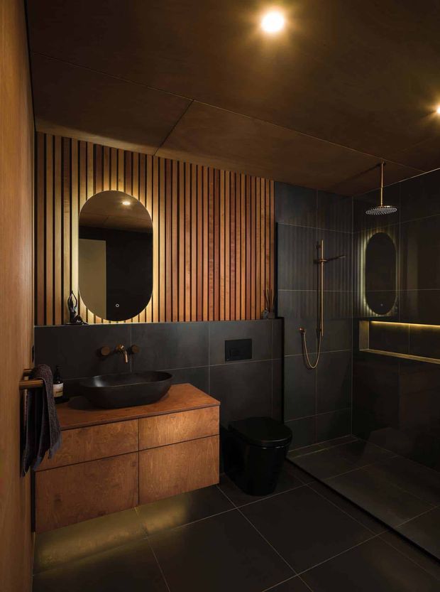 A moody contemporary bathroom with a floating timber vanity, dark vessel basin, matte black toilet and dark tiled walls and floor. A vertical timber slat feature wall frames an arched mirror, while a walk-in shower with brass fixtures and a second arched mirror occupies the right side. Warm concealed and ceiling lighting creates an intimate, luxurious atmosphere.