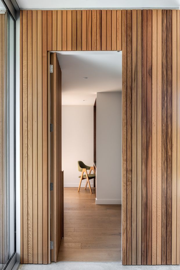 A contemporary interior entry view framed by a full-height vertical timber slat wall and matching concealed or flush timber door. The open doorway reveals a light neutral hallway with timber flooring, recessed ceiling lights, white walls, and a mid-century style chair in the background. Floor-to-ceiling glazing is visible along the left edge.