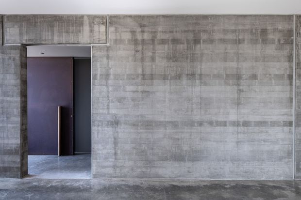 Minimalist interior architectural view dominated by a raw, mottled grey concrete wall and matching concrete floor. A recessed doorway at left contains a dark timber or painted door with a slim vertical metal handle, creating a stark, monolithic composition.