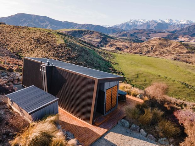 A compact modern cabin or outbuilding set into a rugged alpine landscape. The building has dark vertically ribbed cladding, a low-pitched or flat roof, timber-framed glazing and a small timber deck, surrounded by native grasses, gravel and rocky terrain. Snow-capped mountains and rolling hills form the background.