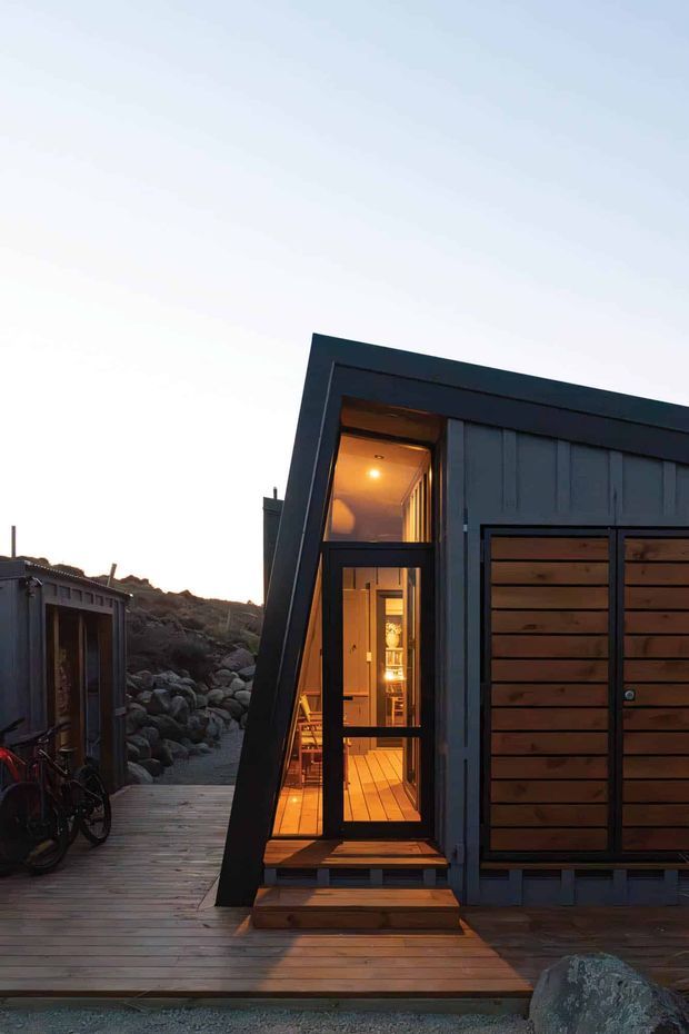Exterior view of a compact dark-clad cabin or small dwelling at dusk, with a strongly angled facade and glazed entry door revealing a warmly lit timber interior. Horizontal timber garage or storage doors occupy the right side, while a timber deck and walkway connect to a second small structure on the left. Rocky terrain and sparse coastal-looking landscape surround the buildings.