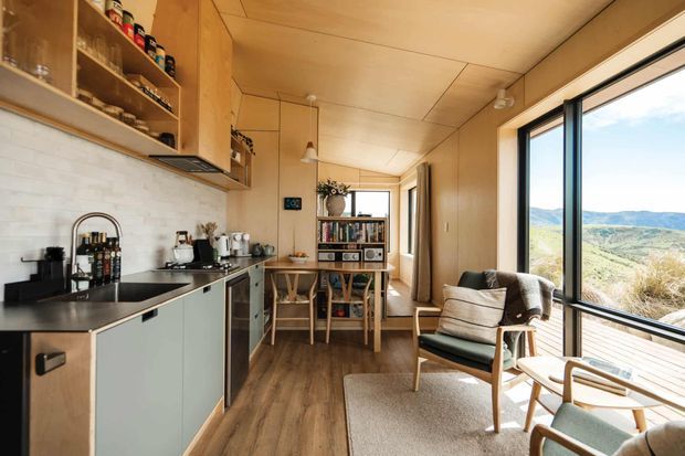 Compact, warm timber-lined open-plan interior combining a small kitchen with a bright living area. The kitchen has pale flat-panel cabinetry, a dark benchtop, open timber shelving and a white tiled splashback. A timber-framed lounge chair, small coffee table, shelving unit and rug sit beside expansive black-framed windows overlooking a rugged natural landscape.