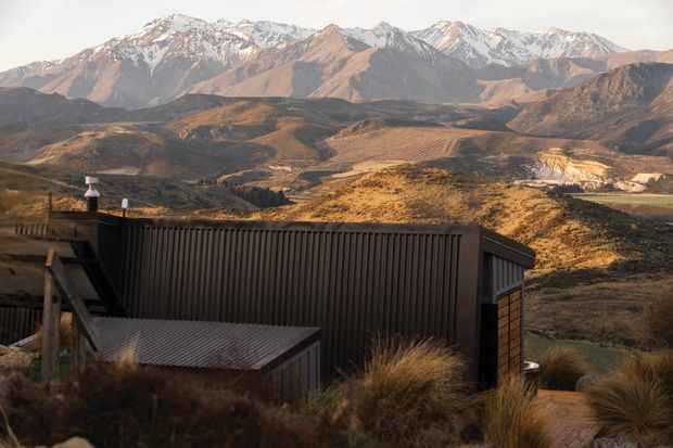 A dark corrugated-metal mountain house or lodge sits in the foreground, partially embedded in a tussock-covered hillside. The view looks across a rugged rural valley with patterned agricultural land toward a dramatic range of snow-capped mountains. The building has a strong angular roofline, vertical ribbed cladding, and warm timber detailing, photographed in soft late-day light.