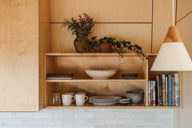 Warm, close-up view of a built-in timber shelving and cabinetry unit. Open shelves display books, ceramics, plates, bowls, folded textiles and potted greenery, with a pendant light at right. A white rectangular tile backsplash runs along the lower portion of the image. The composition has a simple, natural, highly crafted interior character.