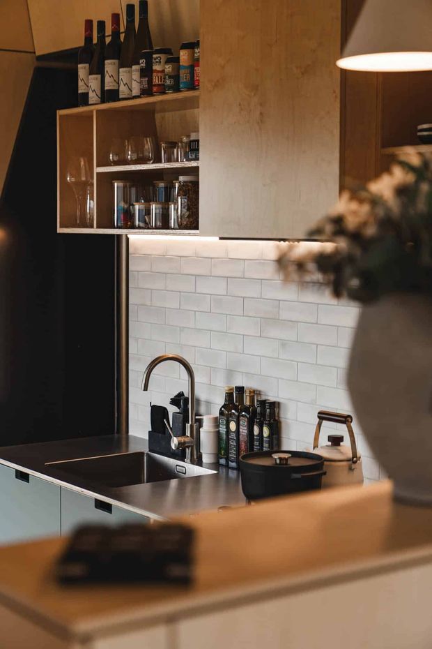 Warmly lit compact kitchen with dark cabinetry, a stainless-steel sink and gooseneck tap, pale subway-tile splashback, open timber shelving displaying wine bottles and glassware, and dark benchtops. A kettle and cookware sit beside the sink, with a softly blurred pendant light and foreground island adding depth.