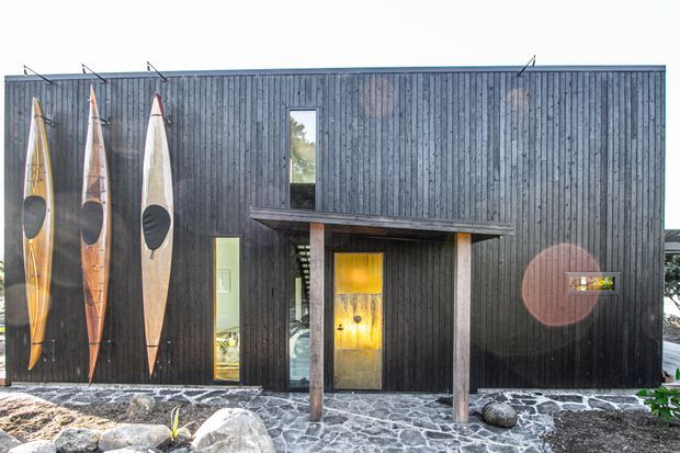 Exterior view of a compact contemporary building clad in dark vertical timber boards. A recessed entry is framed by timber posts and a shallow projecting canopy, with a warm timber door and narrow vertical windows. Several surfboards are mounted vertically against the facade, while irregular dark stone paving and boulders form the foreground.