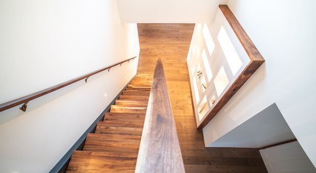 Bright interior view looking up a timber staircase. Warm timber treads, flooring, handrails and a central balustrade contrast with white walls and a white geometric balustrade, with strong natural light entering from above.