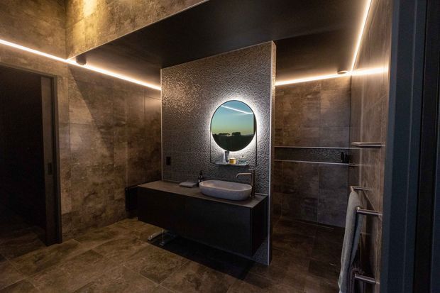 A dark, luxurious contemporary bathroom with large-format stone-look tiled walls and floor, a floating dark vanity with a white vessel basin, a round backlit mirror, concealed linear lighting, and a glass-enclosed shower. A towel rail is visible at right, and the mirror reflects an exterior landscape.