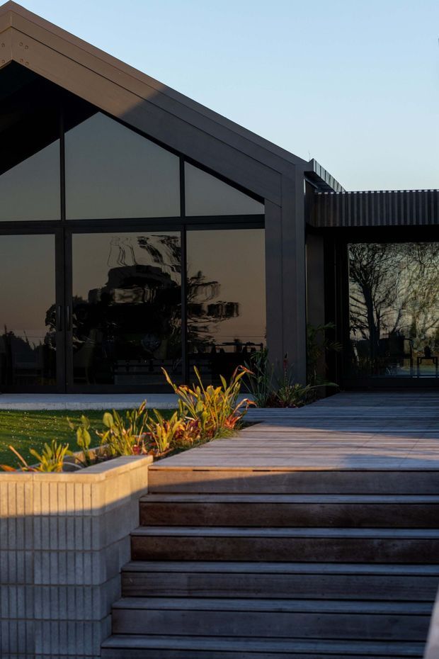 Exterior view of a contemporary single-storey building with a prominent sloping roofline, dark metal-framed floor-to-ceiling glazing, and a flat-roofed wing. Broad timber decking and steps lead up to the entrance, bordered by a built-in rendered planter with linear detailing and ornamental planting. The glazing reflects surrounding trees and landscape.