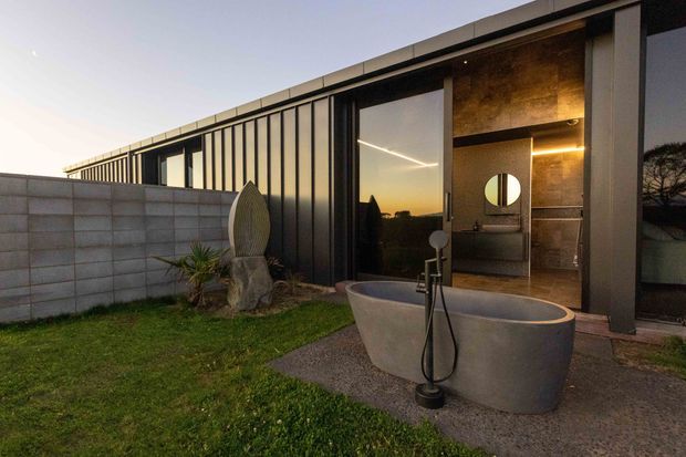 Exterior view of a modern, single-storey house at dusk, featuring a dark metal-framed facade with extensive glazing and a partly enclosed outdoor bathroom area. A freestanding oval bathtub sits on a paved terrace in front of an open bathroom with a round mirror, vanity, and warm recessed lighting. A concrete block boundary wall and landscaped lawn frame the scene.