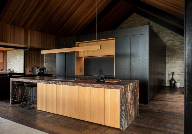 A dramatic, dark-toned contemporary kitchen with a large timber-clad island, a richly patterned stone benchtop and waterfall end, integrated timber cabinetry, dark metal accents, and a timber-lined pitched ceiling. A compact dining or living area is visible beyond the kitchen, with exposed masonry and large glazing contributing to the warm, lodge-like atmosphere.