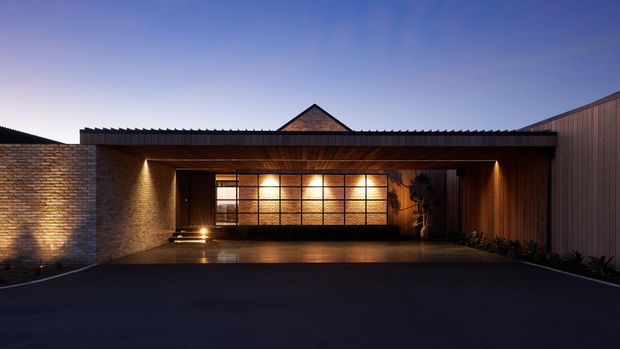A dusk exterior view of a contemporary residential entry or carport, with a broad flat-roofed canopy, warm timber-lined soffit, exposed brick side wall, dark vertical cladding, and a large illuminated gridded glazed doorway or garage door. The paved forecourt dominates the foreground, with subtle planting along the edges and a pitched roof form visible behind.