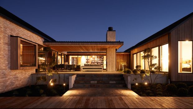 Twilight exterior view of a low, U-shaped contemporary residence arranged around a central courtyard. The home combines warm brick walls, dark vertical timber cladding, extensive illuminated glazing, a broad covered outdoor living area, stepped stone paving, timber decking, landscaped planting, and a prominent chimney. The composition emphasizes indoor-outdoor connection and courtyard living.
