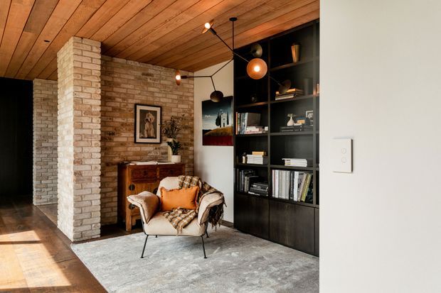 Warm, moody living area featuring exposed light brick walls, a timber-lined ceiling, dark built-in shelving, a vintage leather lounge chair, timber sideboard, artwork, books, and sculptural pendant lighting. The space has an eclectic mid-century character with strong material contrast and a layered, inviting atmosphere.