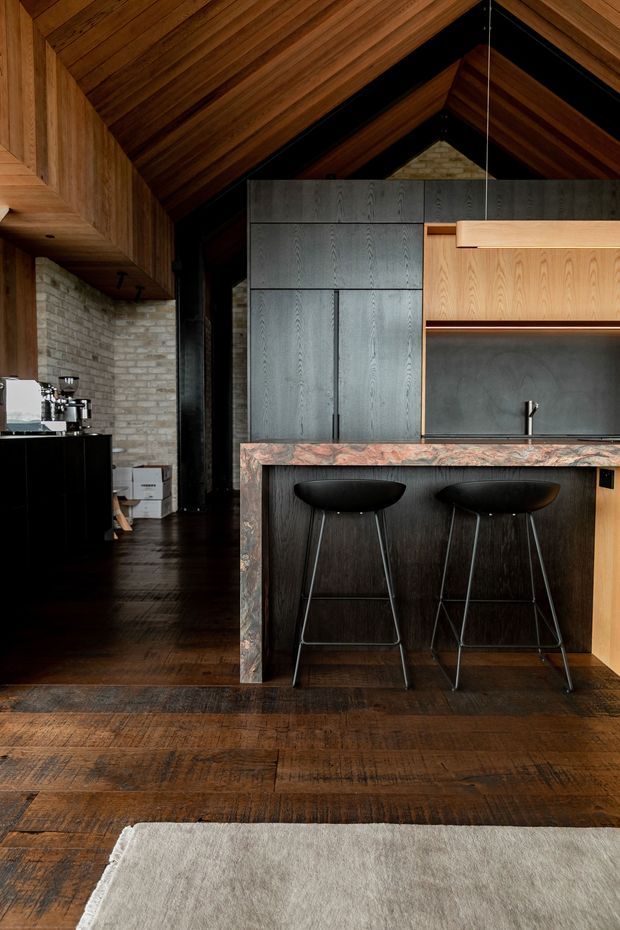 A richly detailed, dark-toned kitchen interior with a dramatic pitched timber-lined ceiling, exposed dark structural framing, warm timber cabinetry, black built-in joinery, a pale brick wall, and a substantial stone island with two black bar stools. The space has dark timber flooring, a linear pendant light, and a refined rustic-modern atmosphere.