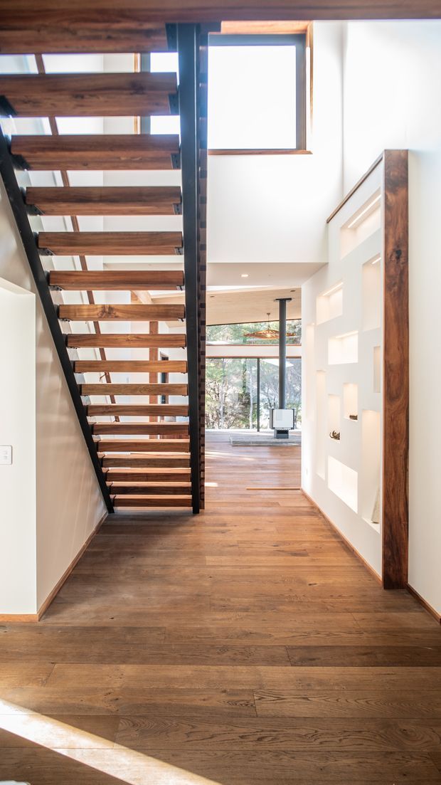 A bright residential hallway and stairway with a floating open-riser staircase featuring dark structural framing and warm timber treads. Wide timber flooring continues through the hall toward a sunlit living area with large glazing. White walls include recessed display niches and a tall timber-framed opening, while a high window and double-height void bring in natural light.