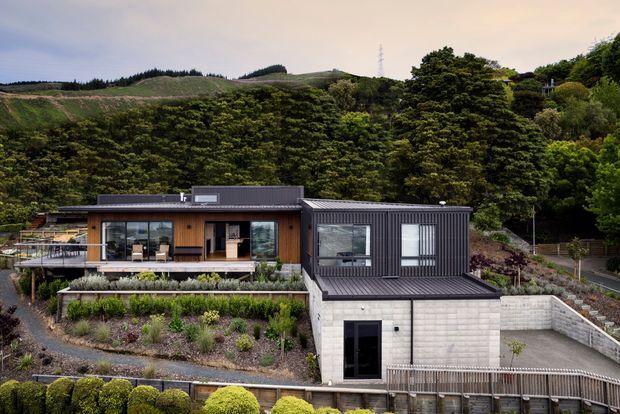 A modern multi-level home set into a landscaped hillside, viewed from above. The building combines dark corrugated cladding and roofing with warm timber wall sections, extensive glazing, a broad terrace, and a lower concrete-block garage or ancillary level. Dense vegetation and rolling wooded hills surround the property.