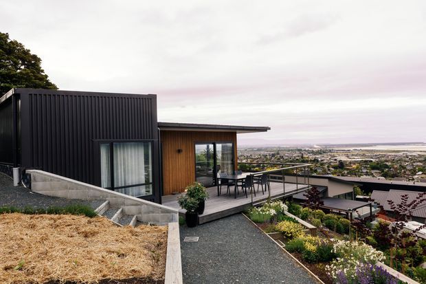 Exterior view of a contemporary split-level home on a sloped site, with dark vertical cladding, warm timber wall panels, broad glazing, and a roof terrace or balcony overlooking a suburban coastal landscape. Gravel paths, planted garden beds, concrete retaining walls, and outdoor dining furniture are visible.