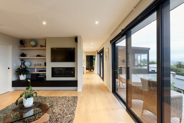 A bright, contemporary open-plan living room with a long circulation corridor and extensive floor-to-ceiling glazing along one side. The room features pale timber flooring, a white ceiling with recessed downlights, a built-in dark media and fireplace wall, open display shelving, a glass coffee table, a textured rug, and indoor plants. The glazing opens to a timber deck with wicker outdoor seating and views toward the coast or water beyond.