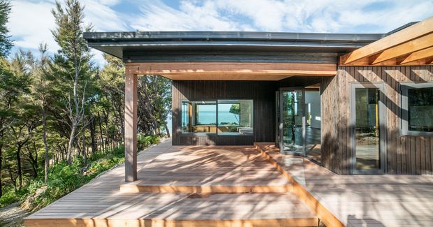 Exterior view of a contemporary single-storey timber-clad house with a broad covered terrace and warm exposed timber beams. Large glazed openings connect the deck to the interior, while the home sits among mature trees on a wooded site. The flat roof and strong indoor-outdoor connection create a calm, natural architectural character.